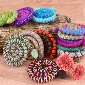 May include: A collection of colorful, hand-felted wool spiral brooches. The brooches are made with different colors of wool and are decorated with embroidery. The brooches are displayed on a piece of driftwood.