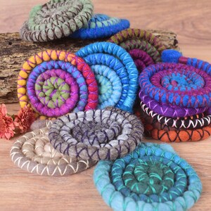 Op de afbeelding: Een verzameling kleurrijke, handgevoelde wollen onderzetters in een spiraalvormig ontwerp. De onderzetters zijn gerangschikt op een houten oppervlak en hebben een verscheidenheid aan kleuren, waaronder blauw, groen, paars en oranje.