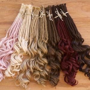 May include: Six strands of synthetic dreadlocks in various colors, including pink, blonde, brown, dark brown, burgundy, and black. The dreadlocks are curly and arranged in a fan shape on a wooden surface.