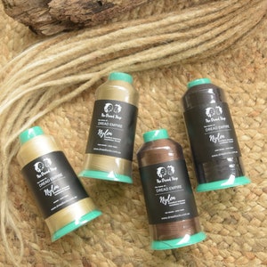 Op de afbeelding: Vier spoelen nylondraad in verschillende bruin- en beigetinten, met groene bases en zwarte etiketten. De etiketten lezen "Dread Empire Nylon". De spoelen liggen op een geweven oppervlak met dreadlock-extensions.