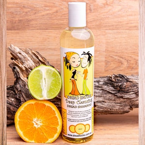 May include: A clear plastic bottle of Dread Empire Deep Cleansing Dread Shampoo with a cartoon illustration of a couple with dreadlocks on the label. The bottle is sitting on a wooden surface with a lime and an orange.