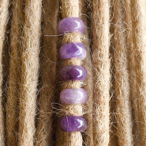 Könnte beinhalten: Nahaufnahme von Dreadlocks mit violetten Amethyst-Perlen. Die Dreadlocks sind hellbraun und die Perlen sind rund und glatt.