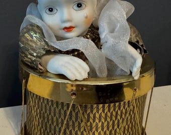 May include: A porcelain clown doll with a painted face and a single teardrop, wearing a gold and black patterned hat and collar. The doll is positioned inside a cylindrical drum-shaped container featuring a gold and black diamond pattern.