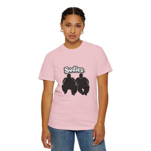 Può includere: T-shirt rosa chiaro con la scritta "Sodies" sopra una grafica nera di due lottatori di sumo. La maglietta è realizzata in un materiale morbido ed è adatta per l'abbigliamento casual.