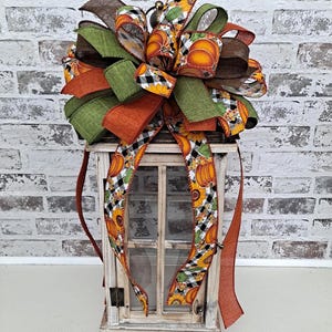 May include: A decorative lantern topped with a large, multi-layered bow made of wired ribbons. The ribbons feature patterns of orange pumpkins, yellow sunflowers, and black and white buffalo check. The lantern has a distressed white wooden frame.