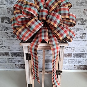 May include: A large, multi-looped decorative bow made of wired ribbon featuring a plaid pattern in shades of red, light blue, brown, and yellow. The bow is positioned on top of a rectangular, light-colored wooden lantern with glass panels.