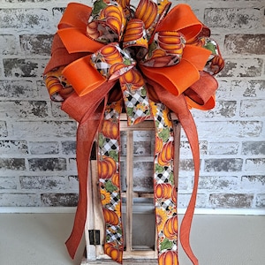 May include: A decorative bow featuring orange burlap ribbon and a patterned ribbon with pumpkins, sunflowers, and black and white buffalo check. The bow is tied with long, trailing ribbon tails that drape down the front of a wooden lantern frame.