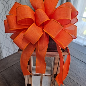May include: A decorative wooden lantern with a large orange bow. The bow is made of a textured ribbon and is tied on top of the lantern. The lantern has a rustic, weathered appearance and features glass panels.
