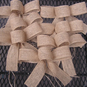 May include: A set of five burlap bows, tied with twine, arranged on a black metal surface. The bows are a light brown color and have a rustic, natural look.