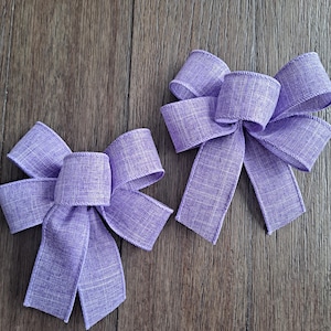 May include: Two lavender-colored fabric bows with a textured finish. The bows are tied with a simple knot and have a rustic, handmade look.