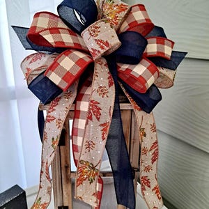 May include: A decorative bow featuring a mix of ribbons in navy blue, red and white buffalo plaid, and a tan burlap-style fabric printed with colorful autumn maple leaves. The bow is arranged with multiple loops and long, trailing ribbon tails.