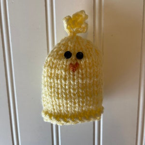 May include: A small knitted chick with black eyes and an orange beak. The chick is yellow and has a small tuft of yarn on top of its head.
