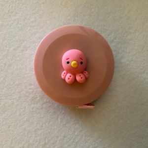 May include: A pink retractable tape measure with a pink octopus design. The octopus has a yellow beak and black eyes.