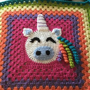 May include: A colorful, crocheted blanket featuring a unicorn design. The blanket has a rainbow gradient background with a white unicorn head, blue nose, black eyes, and a spiraled horn. The unicorn's mane is made of colorful yarn.