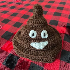 May include: A brown crocheted hat shaped like a pile of poop with a white smiling face and two eyes.