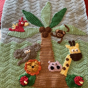 May include: A crocheted baby blanket featuring a green background with a brown palm tree in the centre. The palm tree has green leaves and is surrounded by crocheted animals, including a red bird, a monkey, a grey elephant, a lion, a purple hippopotamus, and a giraffe.