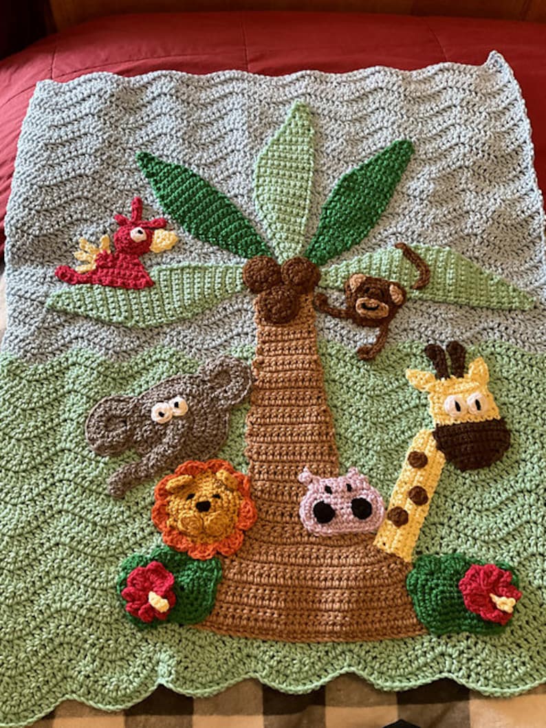 May include: A crocheted baby blanket featuring a green background with a brown palm tree in the centre. The palm tree has green leaves and is surrounded by crocheted animals, including a red bird, a monkey, a grey elephant, a lion, a purple hippopotamus, and a giraffe.
