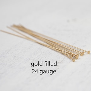 May include: A collection of gold-filled head pins, 24 gauge, arranged on a light-colored surface. The pins have a straight wire shaft and a flat, round head. The text "gold filled" and "24 gauge" is visible.