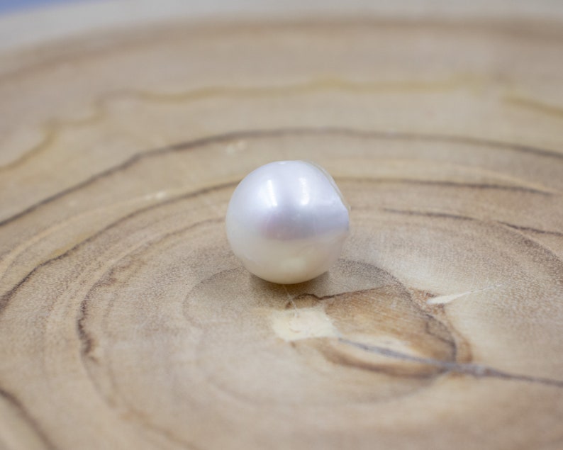 Pu&ograve; includere: Una perla bianca e lucente con una superficie liscia. La perla rotonda riflette la luce, su uno sfondo marrone chiaro con venature del legno. L'immagine presenta un'elegante perla.