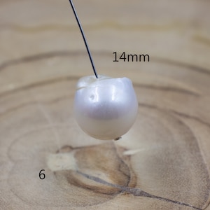 Pu&ograve; includere: Una perla bianco-avorio, di circa 14 mm di diametro, &egrave; forata da un filo sottile. La perla ha una forma leggermente irregolare e una superficie liscia e riflettente. Il numero "6" &egrave; visibile nell'angolo in basso a sinistra.