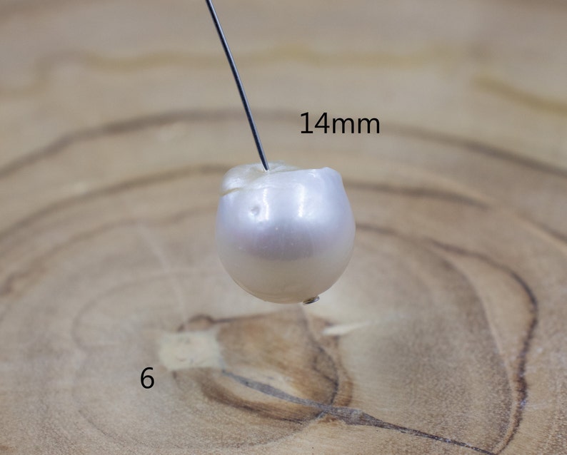 Pu&ograve; includere: Una perla bianco-avorio, di circa 14 mm di diametro, &egrave; forata da un filo sottile. La perla ha una forma leggermente irregolare e una superficie liscia e riflettente. Il numero "6" &egrave; visibile nell'angolo in basso a sinistra.