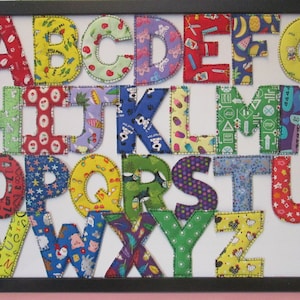 May include: A framed alphabet wall art with each letter made from colorful fabric scraps. The letters are sewn together with a zig-zag stitch and are displayed on a white background.