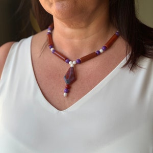 May include: A necklace featuring brown, white and purple beads. The necklace has a pendant made from a reddish-purple stone with white and grey markings.