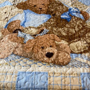 May include: A quilted baby blanket with a brown teddy bear design. The blanket features a blue and beige checkered pattern with a blue and white striped ribbon on the teddy bear.