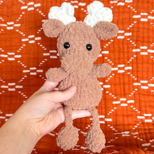 May include: A brown crocheted moose toy with white antlers. The moose is sitting on a red and white patterned blanket.