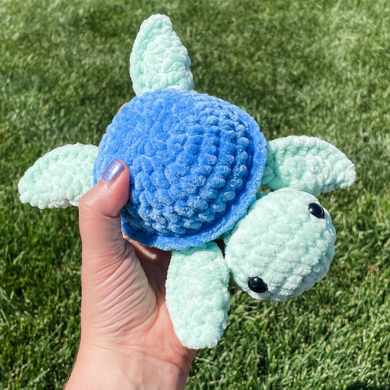 May include: A crocheted sea turtle toy with a blue shell and light green limbs. The turtle has black eyes and is held in a hand.