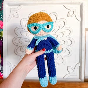 May include: A handmade crochet superhero doll with a light brown head of hair and blue-rimmed glasses. The doll is wearing a dark blue suit with light blue accents and a cape. The doll is approximately 10 inches tall.