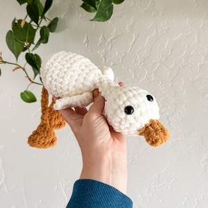 May include: A white crocheted duck with brown felt feet and a beak. The duck is being held by a hand.