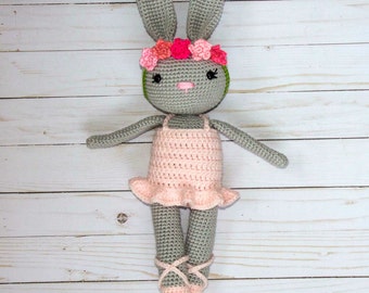 Puede incluir: Un conejito de crochet gris que lleva un vestido rosa y una corona de flores con detalles rosas y verdes. El conejito lleva zapatos de ballet rosas.