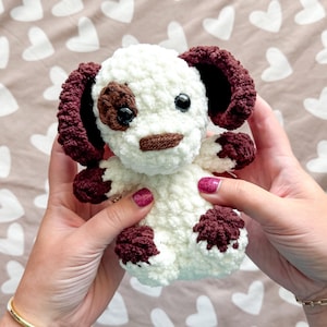 May include: A handmade crochet dog plushie with a white body, brown ears, and brown spots. The dog has black safety eyes and a brown nose. The plushie is held in front of a heart-patterned background.