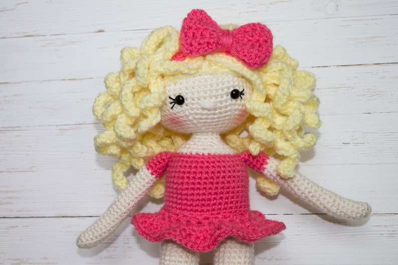 Pu&ograve; includere: Bambola all'uncinetto fatta a mano con capelli biondi ricci e un fiocco rosa. La bambola ha un viso bianco con occhi neri e guance rosee. Indossa un vestito rosa con gonna a balze e maniche bianche.