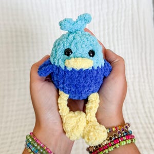 May include: A handmade crochet bird plushie in shades of blue and yellow. The bird has a light blue head with a small crest, a dark blue body, and yellow feet and beak. Black safety eyes complete the design. A whimsical, soft toy.