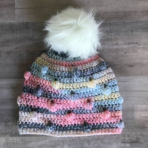 May include: A colorful crocheted beanie with a white pom pom. The beanie is made with a variety of colors, including pink, blue, yellow, and gray. The beanie has a textured design with small bumps throughout.