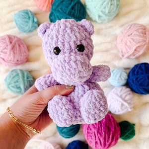 Puede incluir: Un juguete de peluche de hipopótamo de crochet morado. El juguete está sentado en una mano y rodeado de ovillos de lana de varios colores.