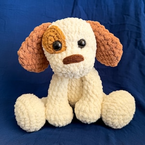 May include: A handmade crochet dog plushie with a white body, brown ears, and a brown spot over one eye. The dog has black button eyes and a brown nose. The plushie is sitting on a blue background.