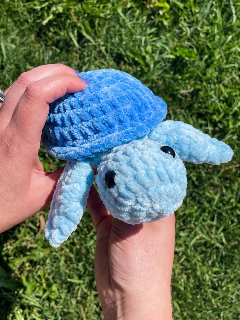 May include: A blue crocheted turtle plush toy with black eyes. The toy is being held by two hands.
