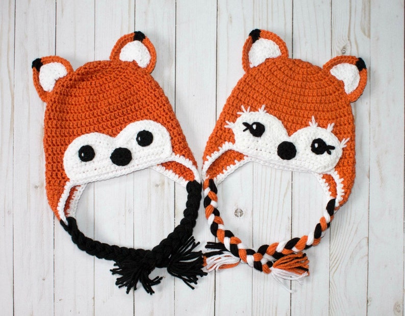 Puede incluir: Dos gorros de zorro de punto de ganchillo naranja y blanco con lazos trenzados negros y blancos. Los gorros tienen ojos negros y una nariz negra.
