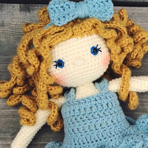 Pu&ograve; includere: Bambola all'uncinetto fatta a mano con viso e braccia color crema e un vestito azzurro. La bambola ha capelli ricci dorati, occhi blu e un fiocco blu abbinato. Le guance sono leggermente arrossate, su sfondo di legno grigio.