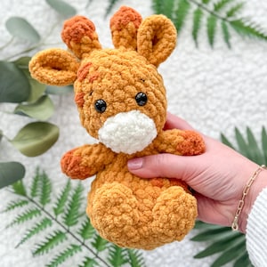 Puede incluir: Un juguete de peluche de jirafa hecho a crochet con una cara blanca y ojos negros. La jirafa es de color marrón claro con detalles naranjas.