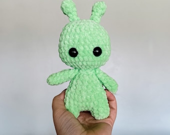May include: A green crocheted plush toy with large black eyes. The toy is shaped like a humanoid figure with two arms and two legs.