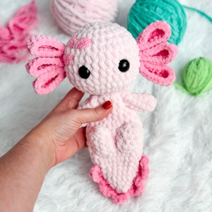 May include: Pink crocheted axolotl plush toy with black eyes and pink frilly gills. The toy is sitting on a white fluffy surface.