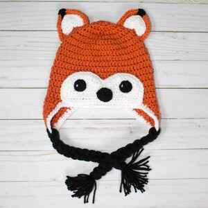 Puede incluir: Un gorro de crochet de zorro con piel naranja y blanca, orejas negras y un cord&oacute;n trenzado negro con borlas.