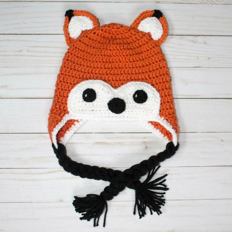 Puede incluir: Un gorro de crochet de zorro con piel naranja y blanca, orejas negras y un cord&oacute;n trenzado negro con borlas.