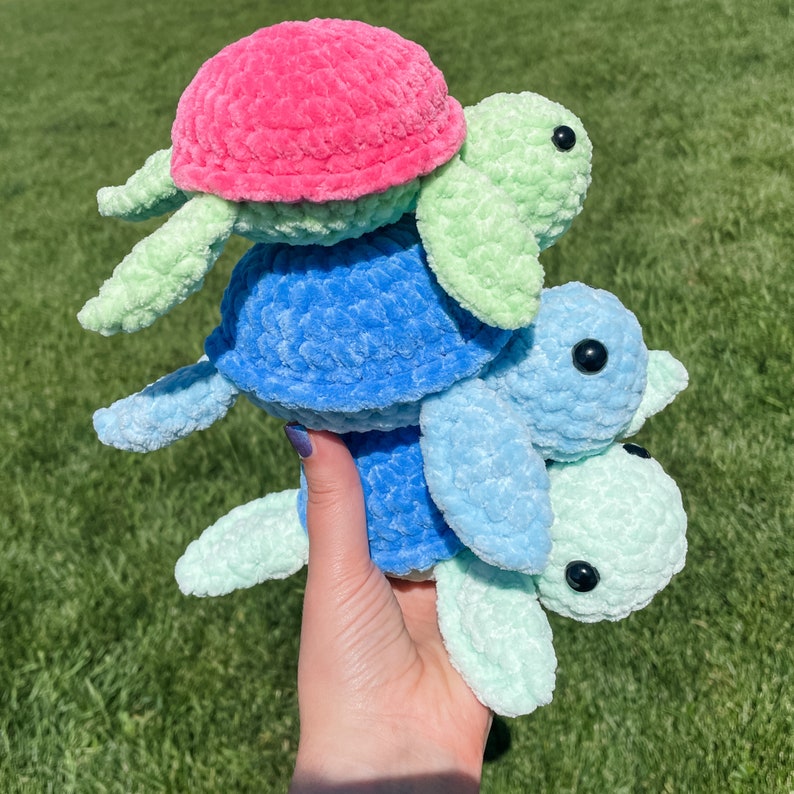 May include: Three crocheted sea turtle toys stacked on top of each other. The top turtle is pink, the middle turtle is blue, and the bottom turtle is light green.