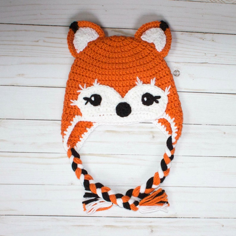 Puede incluir: Sombrero de zorro de punto de ganchillo naranja y blanco con lazos trenzados negros y naranja.