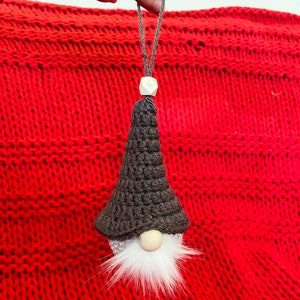 May include: A black and white gnome ornament with a white beard and a wooden bead nose. The gnome is hanging from a red knitted background.